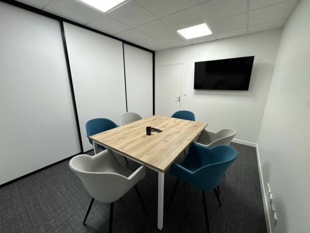 Salle de réunion moderne équipée d’une table en bois clair entourée de fauteuils bleus et gris, avec un écran mural et des placards intégrés aux portes coulissantes blanches.